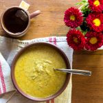 A bowl of broccoli soup with a cup of hot tea and some flowers.