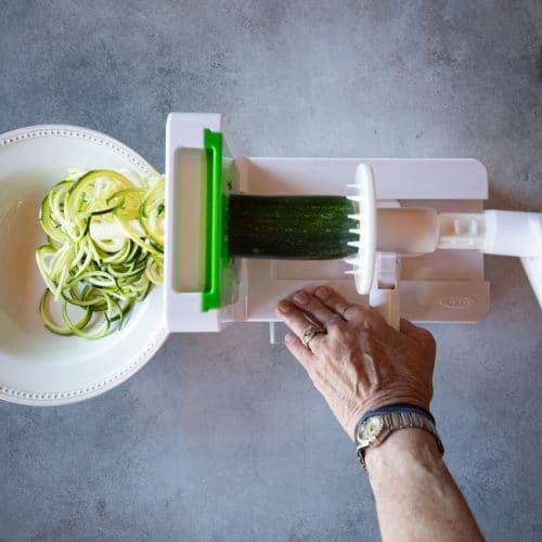 A zucchini positioned in a spiralizer between the grater and the handle. Some zoodles are spilling into a bowl.