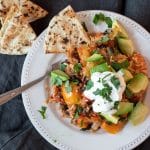A plate of stuffed pepper casserole, Mexican style, topped with parsley, avocado and sour cream, with pita bread on the side.