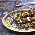 Serving plate with zoodles and grilled veggies skewers, with a serving spoon and fork.