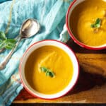 Two bowls of roasted eggplant soup pureed with a tomato, along with two spoons.