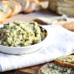 A small bowl of artichoke dip made with green olives and manchego cheese placed on a napkin with slices of bread in the background.
